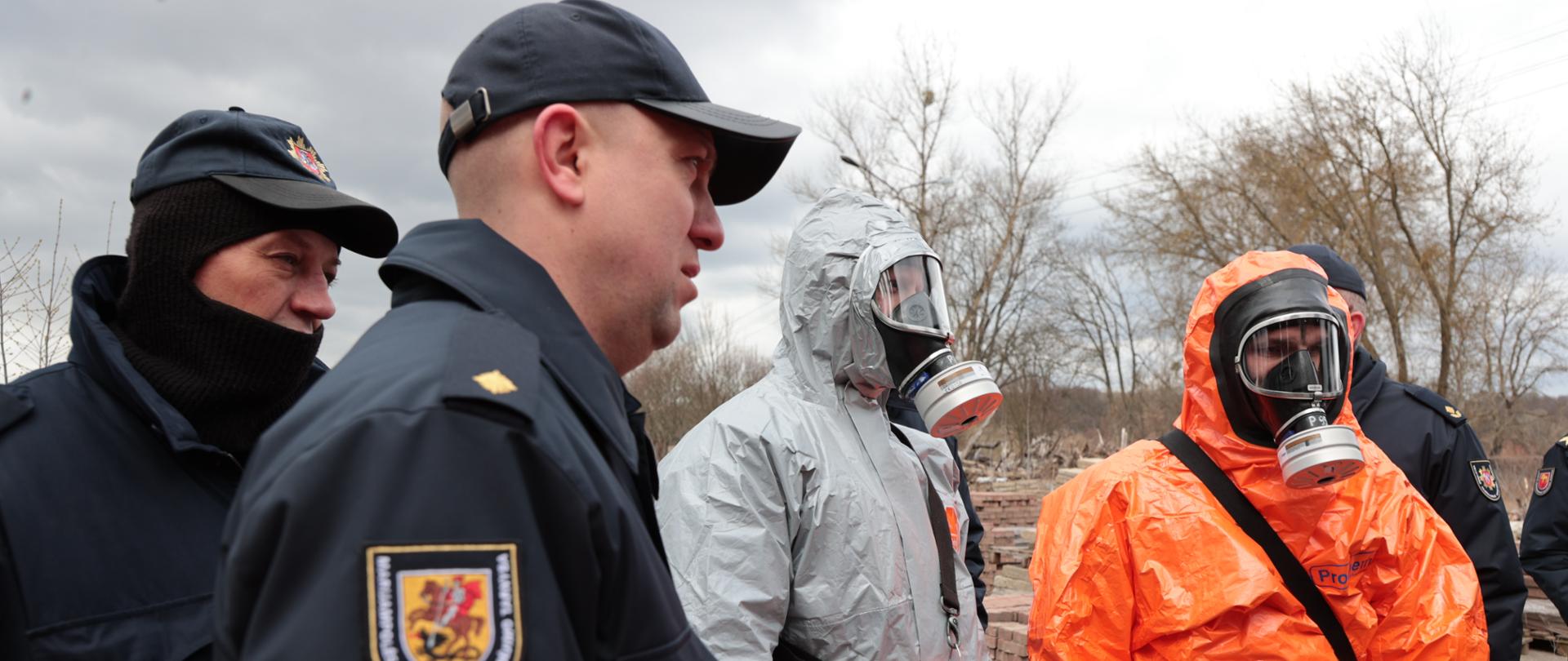 Dwóch ratowników chemicznym w ubraniu specjalnym w kolorze szarym i pomarańczowym oraz maskach z filtropochłaniaczami. Obok nich widoczni są litewscy strażacy w ubraniu sztabowym.