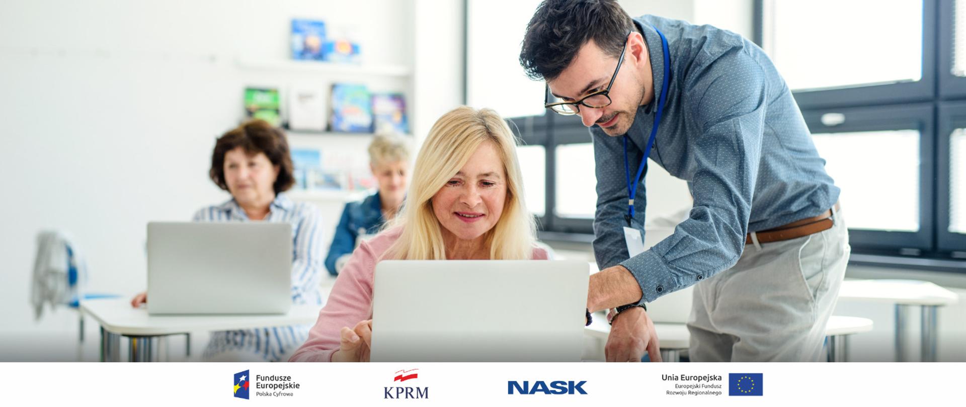 Na zdjęciu nowoczesna sala szkoleniowa, w niej trzy kobiety w wieku 50 plus, korzystające z laptopów. Przy jednej z nich nachyla się młody mężczyzna w okularach i koszuli wskazując coś ręką na jednym z laptopów. 