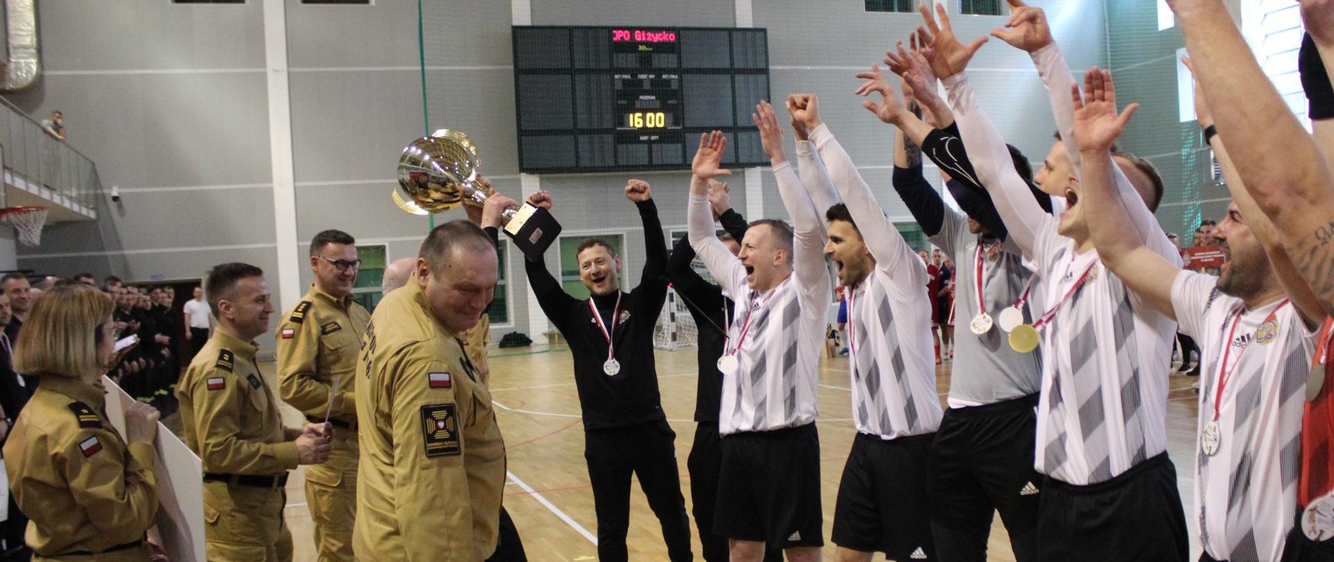 Zwycięska drużyna z KW PSP w Łodzi stojąca w szeregu w trakcie wznoszenia okrzyku radości. 