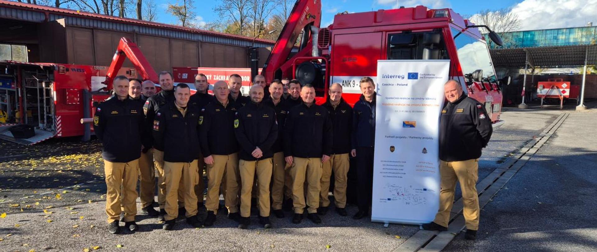 Grupa około szesnastu mężczyzn w strojach służbowych (czarne kurtki, żółte/beżowe spodnie) stoi na zewnątrz przed dużym, czerwonym pojazdem strażackim z ramieniem hydraulicznym. Mężczyźni są uśmiechnięci i patrzą wprost na obiektyw. Na pierwszym planie po prawej stronie znajduje się biały rollup/baner z logo Interreg Czechy-Polska i tekstem w języku polskim dotyczącym projektu. Tło stanowią budynki gospodarcze i inne pojazdy strażackie.