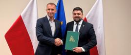 Dwaj mężczyźni pozują do zdjęcia, podając sobie ręce. Mężczyzna stojący po prawej trzyma w rękach zieloną ozdobną teczkę ze złotym logo KRUS. Z tyłu za nimi dwie flagi Polski i flaga Unii Europejskiej.