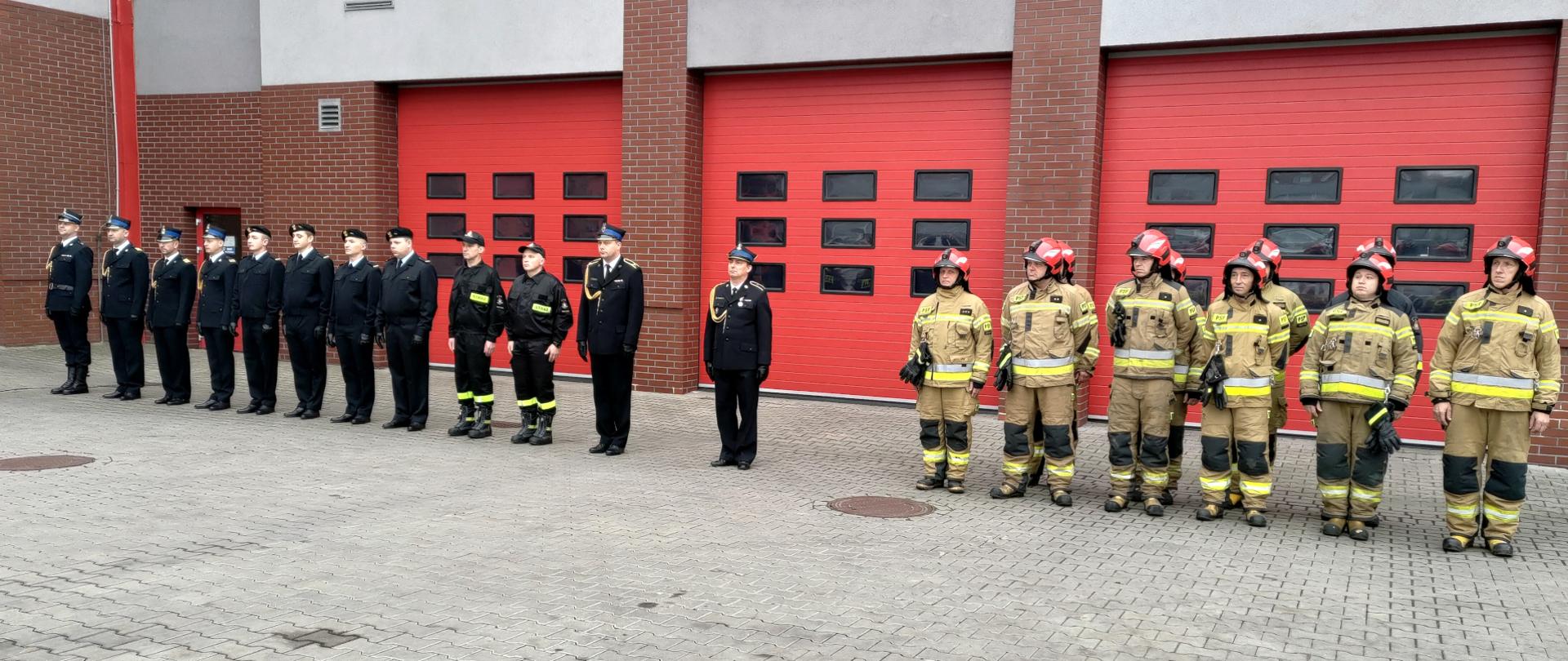 Zdjęcie przedstawia grupę strażaków w umundurowaniu wyjściowym oraz ubraniach specjalnych stojących w dwóch pododdziałach. W tle bramy garażowe.