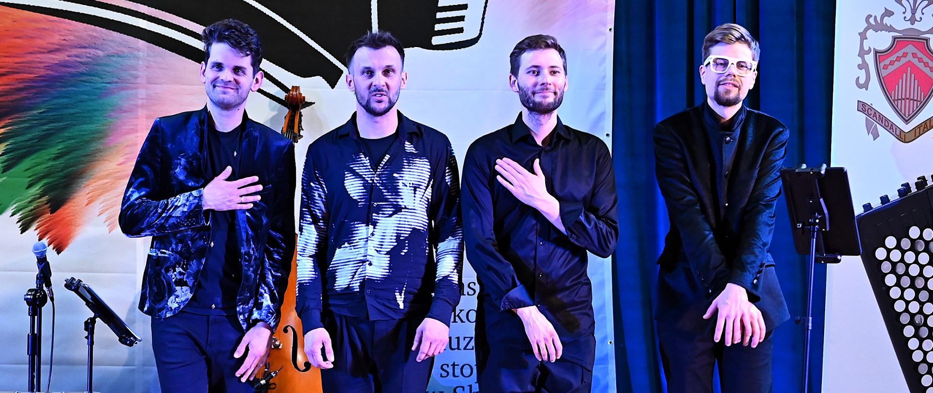 Czterech muzyków stojących na scenie podczas oklasków, wokół nich instrumenty i banery konkursowe