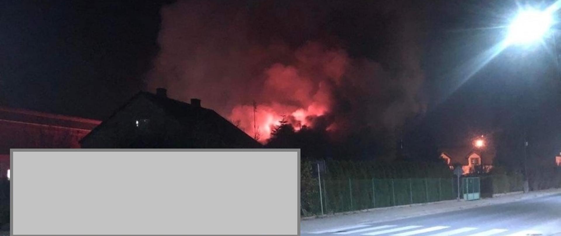 Pora wieczorna, w oddali widać dym oraz płomienie z połaci dachowej budynku mieszkalnego. Na pierwszym palnie droga przebiegająca przy posesji na której znajduje się budynek objęty pożarem, na miejscu nie ma jeszcze służb ratowniczych