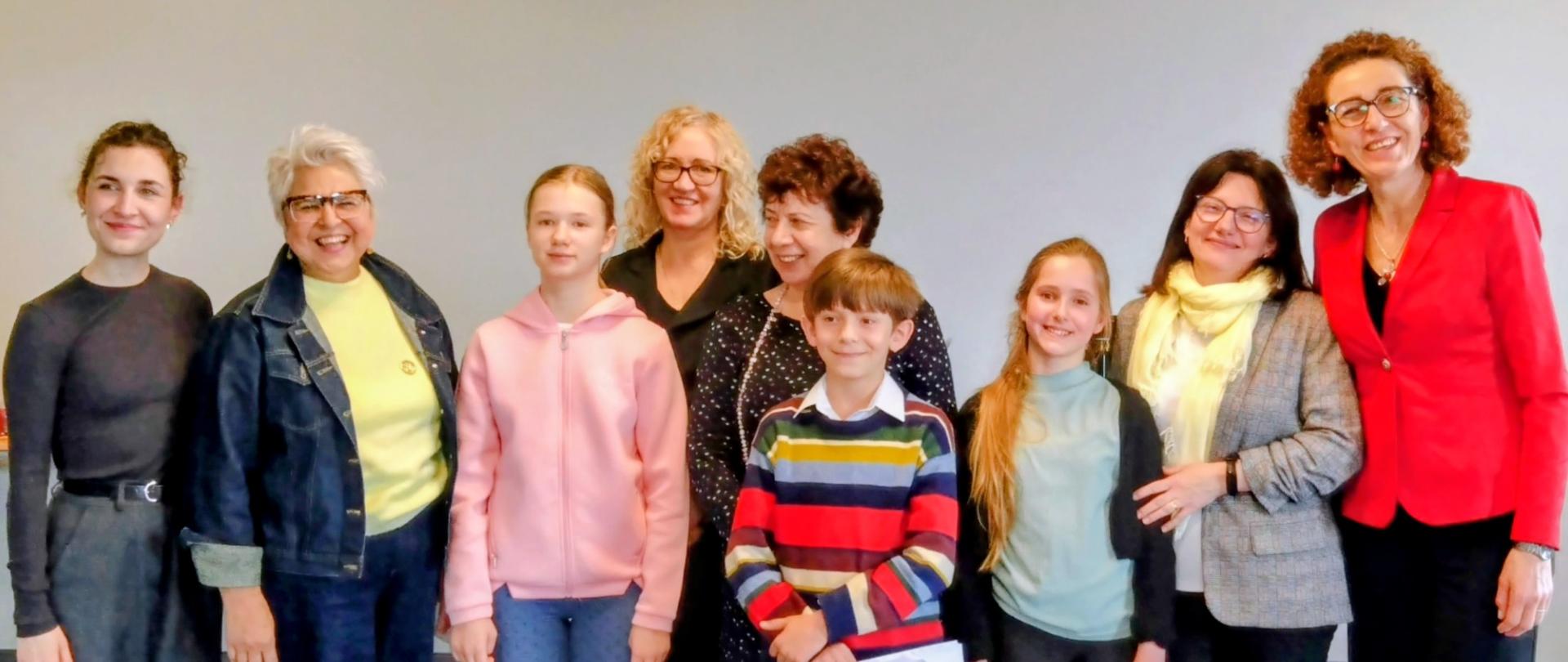 Zdjęcie przedstawia grupę dziewięciu osób pozujących do wspólnej fotografii w jasnym pomieszczeniu na tle neutralnej, szarej ściany. Grupa składa się z dorosłych kobiet oraz dzieci, co sugeruje charakter rodzinny lub szkolny/edukacyjny. Kobieta w czarnym golfie i ciemnych, szerokich spodniach. Kobieta w kurtce jeansowej, jasnożółtej bluzce i ciemnych spodniach. Młoda dziewczyna w różowej bluzie z kapturem i jeansach. Kobieta w okularach z blond, kręconymi włosami, ubrana na czarno (stoi nieco z tyłu). Chłopiec w kolorowym, pasiastym swetrze i jasnych spodniach, trzymający kartki papieru. Kobieta z krótkimi, ciemnymi włosami w ciemnej sukience w drobny wzór (stoi za chłopcem). Dziewczynka w jasnoniebieskiej bluzce, czarnym sweterku i białych butach. Kobieta w okularach z jasnym szalem i szarym żakiecie. Kobieta w wyrazistym, czerwonym żakiecie i ciemnych spodniach. Wszyscy uczestnicy są uśmiechnięci i zwróceni w stronę aparatu. Oświetlenie jest miękkie i równomierne, a kompozycja zdjęcia jest statyczna i oficjalna.