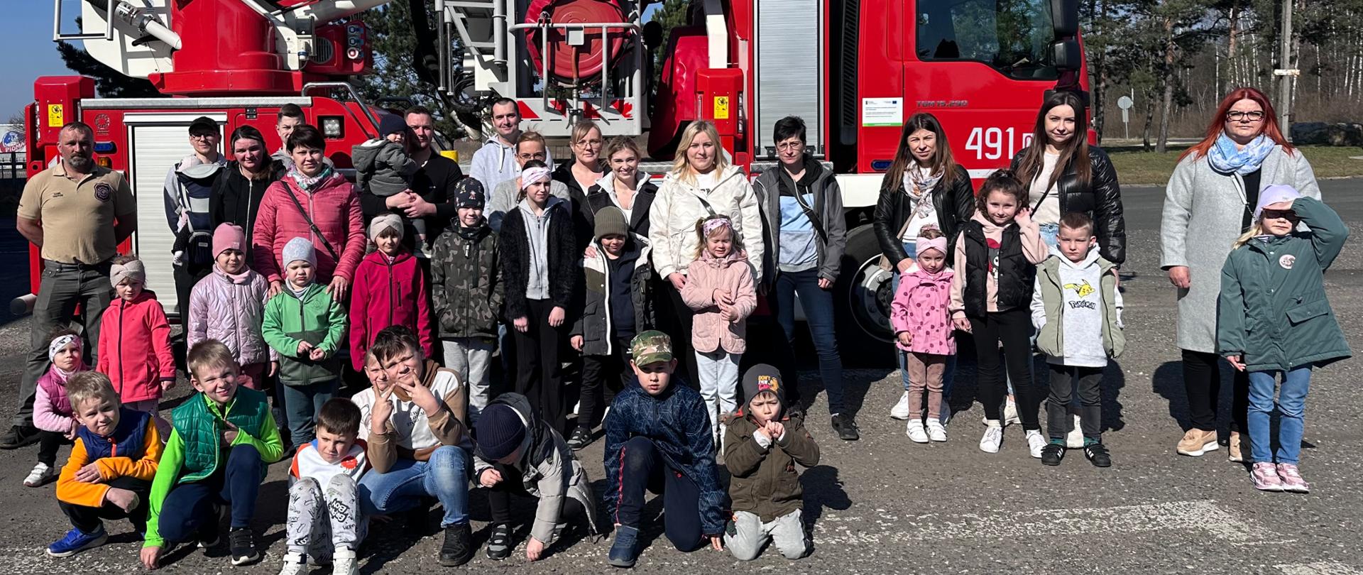 Na zdjęciu znajdują się strażacy w ubraniach służbowych, osoby cywilne i dzieci ustawione do zdjęcia na tle samochodu bojowego