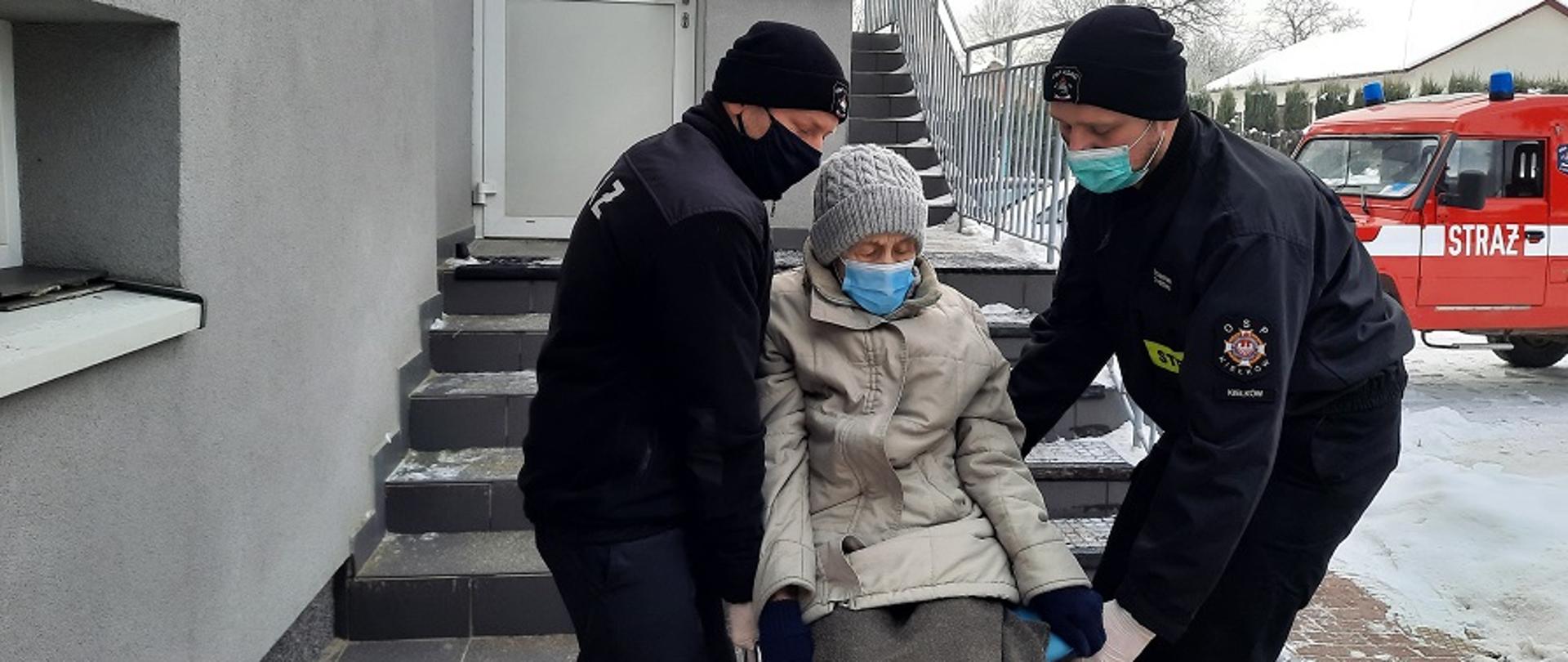 Na zdjęciu widać strażaków niosących starszą kobietę siedzącą nie krześle w kierunku stromych schodów zewnętrznych pokrytych płytkami. Na ścianie budynku widać tablicę „NFZ”, w tle samochód strażacki.