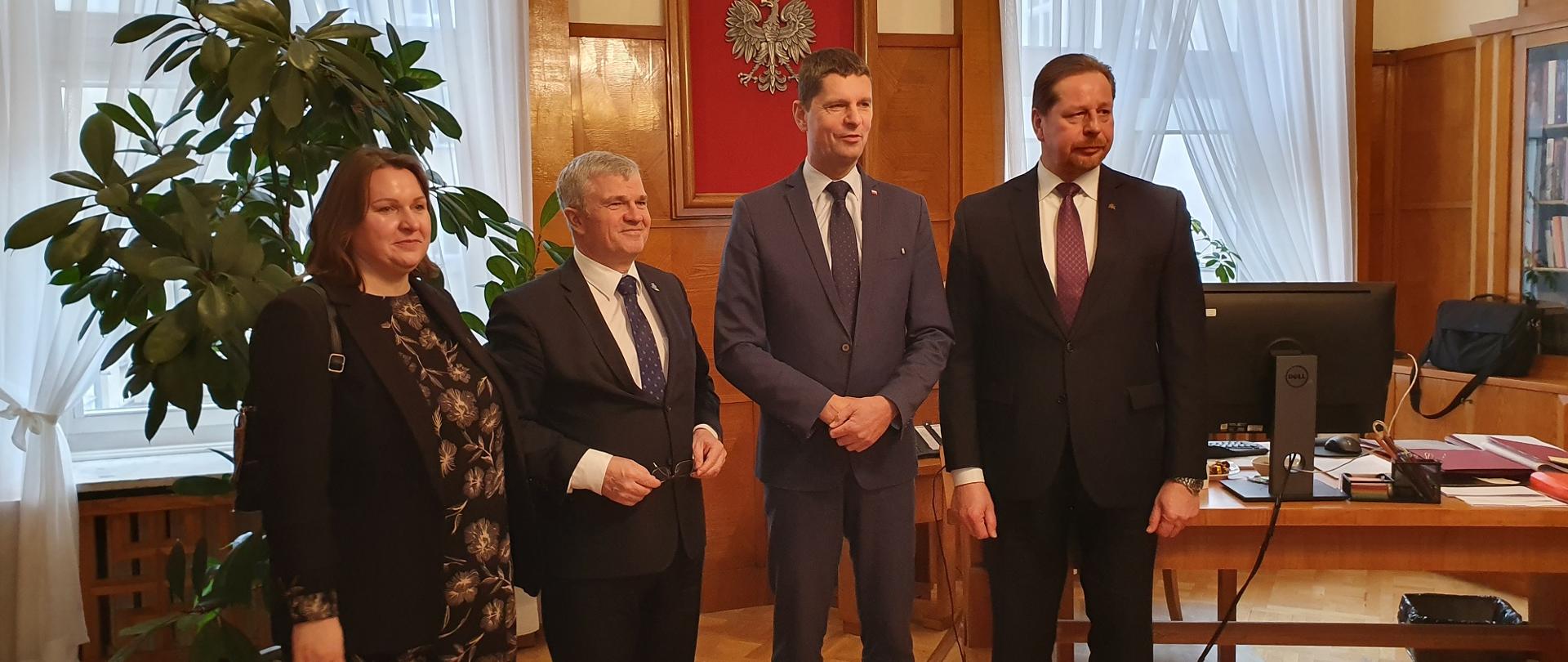 Wiceminister Piontkowski, ubrana na brązowo kobieta i dwóch mężczyzn w garniturach stoją w eleganckim pokoju, za nimi drewniany stolik, duża roślina w doniczce, dalej żółta ściana pokryta drewnianymi panelami na której wisi godło Polski i dwa okna.