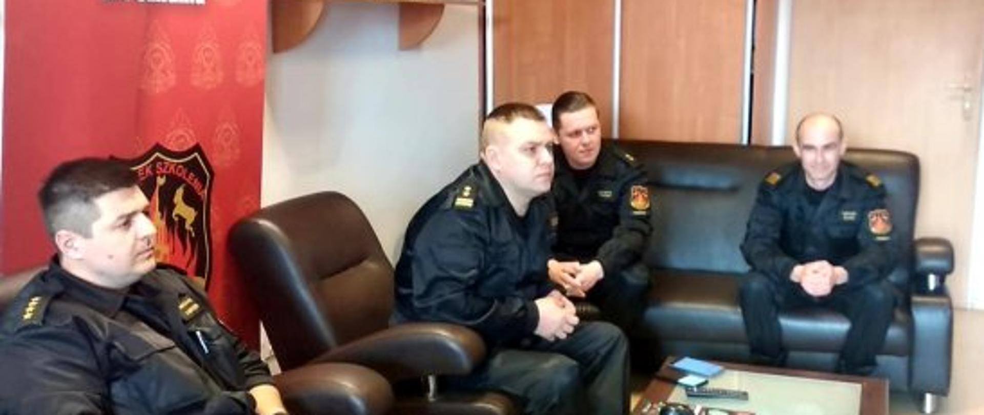 na kanapie siedzą funkcjonariusze, przed nimi stoi niska ława. Patrzą w telewizor , za nimi stoi czerwony baner