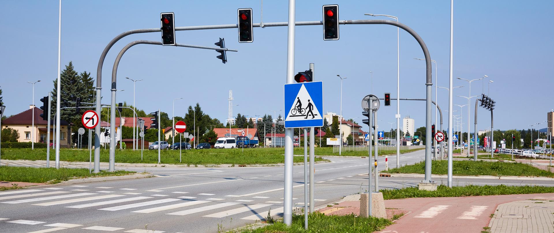 Na zdjęciu pogodna aura, świeci słońce, widać znak drogowy i przejście dla pieszych. Widać także sygnalizatory świetlne. Nie ma ludzi ani samochodów.