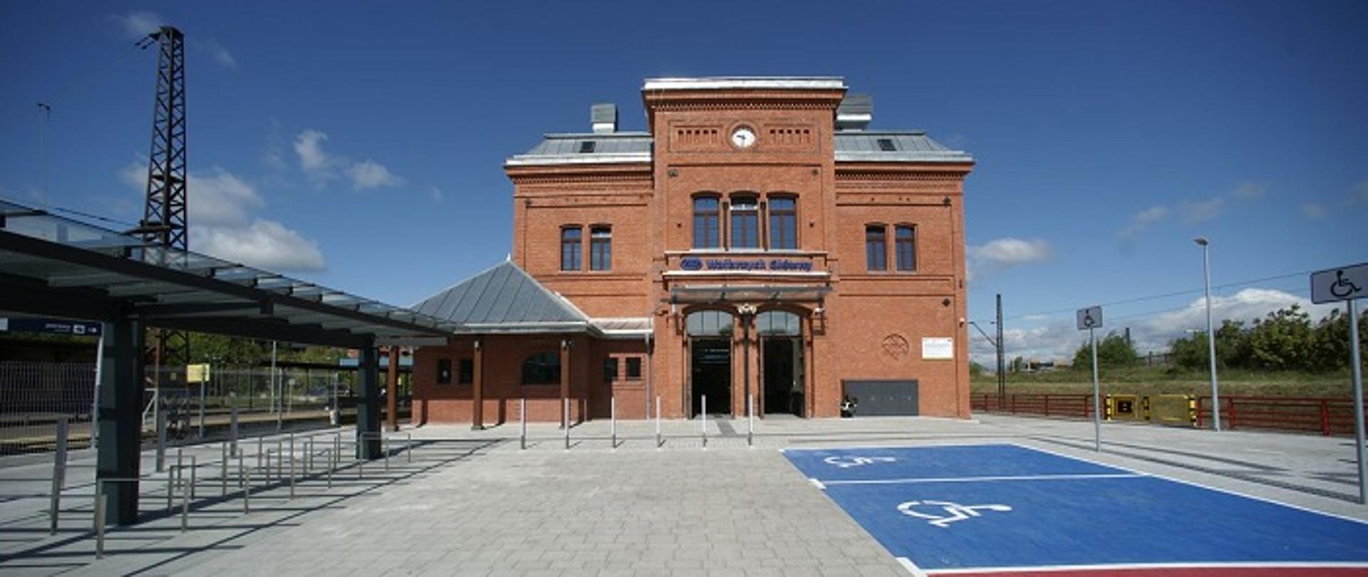 Zdjęcie przedstawia budynek dworca kolejowego z czerwonej cegły. Przed budynkiem widoczny parking z miejscami dla osób z niepełnosprawnością, wiata na rowery oraz wejście na peron.