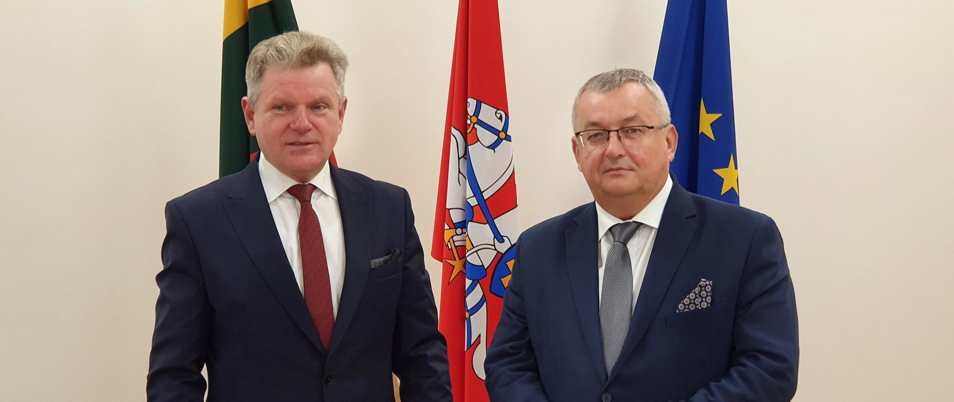 Na zdjęciu widać dwóch mężczyzn, ubranych w ciemne garnitury oraz białe koszule. Pierwszy mężczyzna po lewej stronie ma czerwony krawat, drugi mężczyzna ma krawat szary. W ich tle widać 3 flagi.