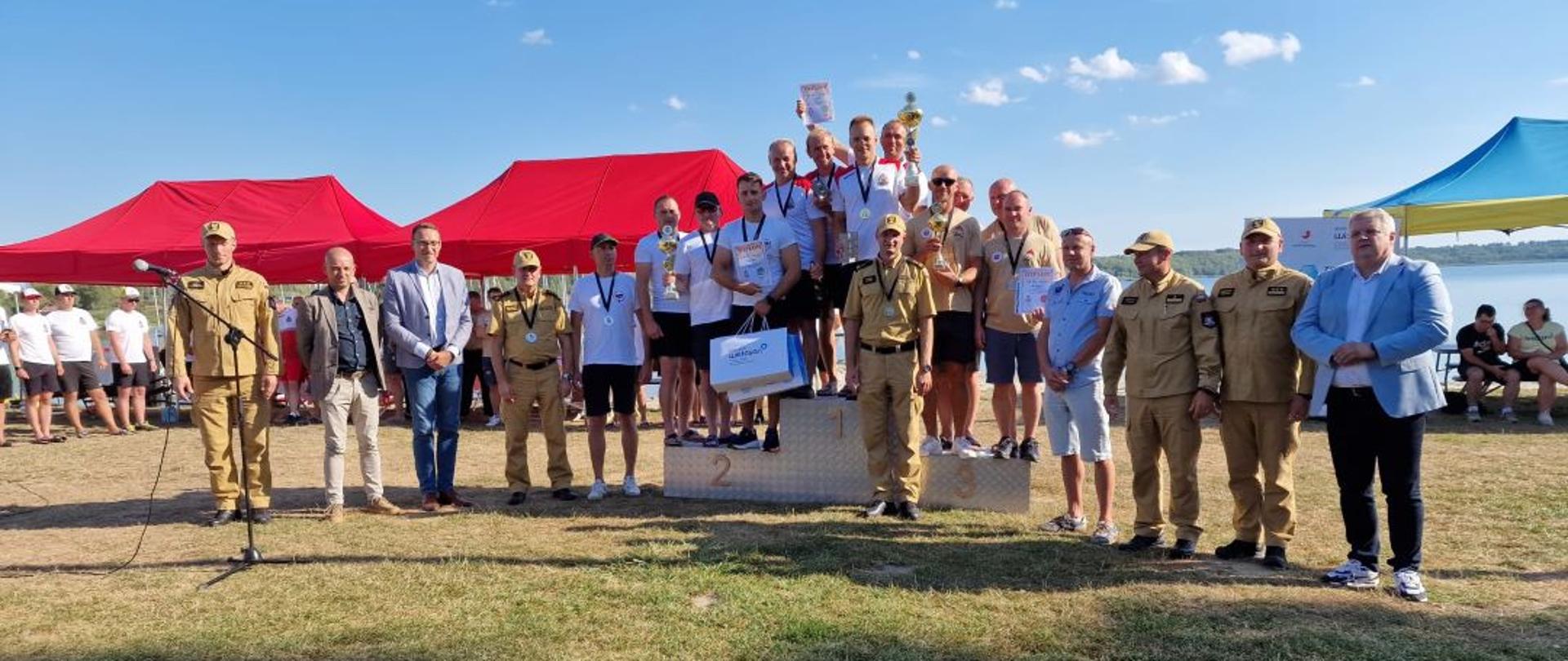 Kolorowa fotografia wykonana w pogodny dzień na zewnątrz na tle Jeziora Tarnobrzeskiego, Przedstawia strażaków płetwonurków na podium zawodów wojewódzkich z wręczonymi dyplomami, medalami i nagrodami ubranych w strojach sportowych i służbowych oraz osoby cywilne stojące obok podium.