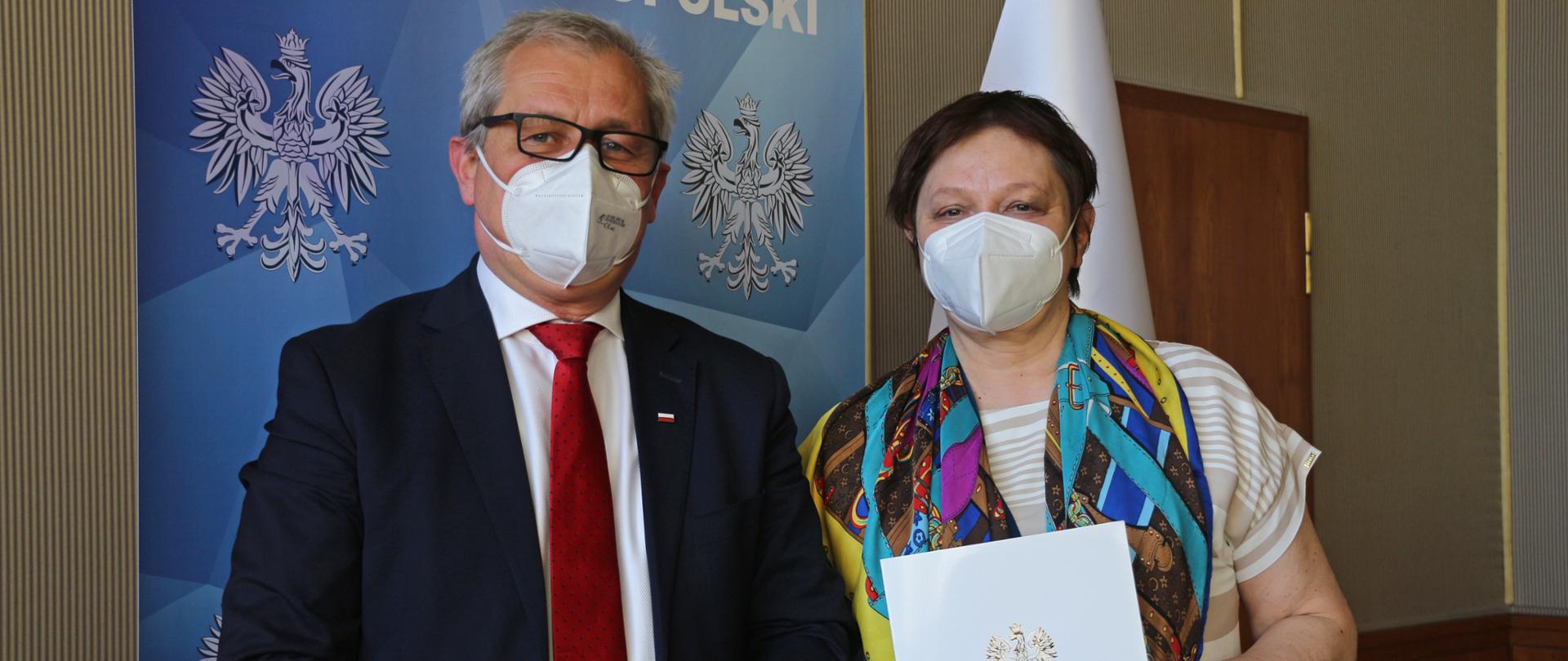 Na zdjęciu po lewej stornie wojewoda opolski oraz - stojąca obok niego, kobieta - pani w średnim wieku, trzymająca w dłoniach białą teczkę z orzełkiem i napisem Wojewoda Opolski. Oboje spoglądają w kadr. Wojewoda na swojej twarzy ma ciemne, korekcyjne okulary. Dłonie pozostawia splecione na wysokości paska. Stojąca obok niego kobieta ma na szyi długi, kolorowy szal. W tle roll up z napisem Wojewoda Opolski oraz wystrój sali, w której odbyło się spotkanie - beżowa ściana.