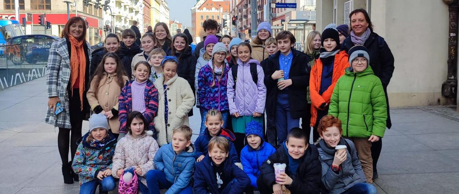 Grupowe zdjęcie wszystkich uczniów biorących udział w wyjeździe uczniów na koncert pt. "Baśń orientalna" we Wrocławiu na tle ulicy oraz budynków w tle 