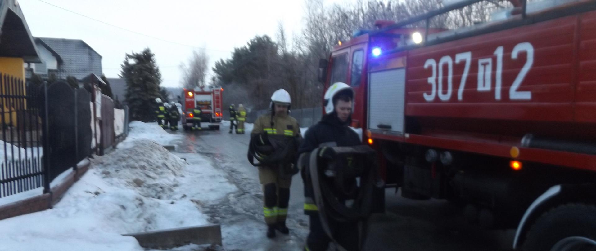 Zdjęcie przedstawia strażaków, którzy ze zwiniętymi wężami idą w kierunku samochodu strażackiego. W tle widać drugi samochód straży pożarnej i grupę strażaków. 