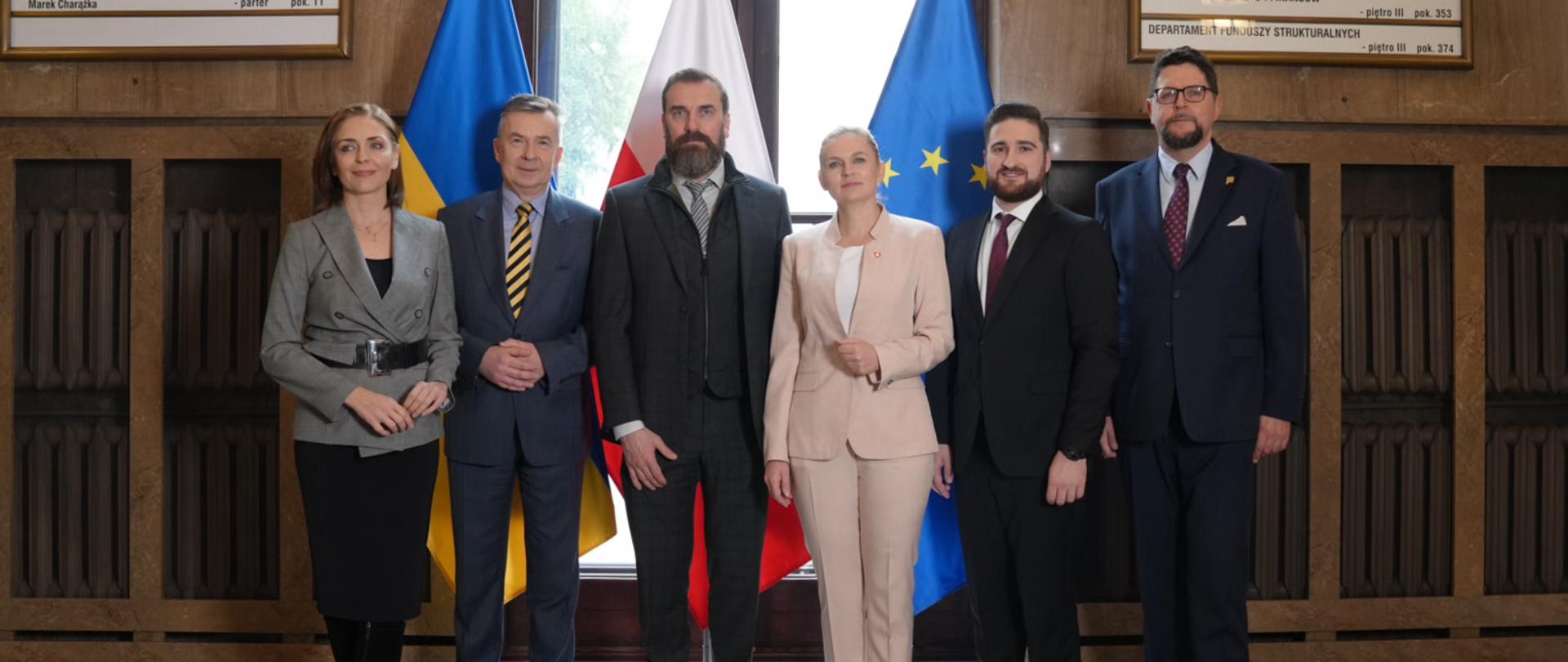 Pod ścianą stoi w rzędzie sześć osób, za nimi okno przed którym stoją flagi Polski, Ukrainy i UE.