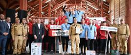 Zdjęcie przedstawia dużą grupę strażaków i organizatorów pozujących do zdjęcia pod drewnianym zadaszeniem. Na pierwszym planie, w centrum, na trzystopniowym podium stoją dwaj mężczyźni w niebieskich koszulkach i dwóch mężczyzn w czerwonych koszulkach, z medalami na szyjach, trzymający w górze puchary i dyplomy. Wokół podium stoją inni mężczyźni w czerwonych i niebieskich koszulkach, również z medalami, a także kilku mężczyzn w garniturach i piaskowych mundurach strażackich. Przed grupą widać nagrody w postaci zapakowanych kartonów. Po prawej stronie, jeden z mężczyzn trzyma tabliczkę z napisem "KM PSP BRZOZOW". W centrum znajduje się również transparent z napisem "KP PSP JAROSŁAW" oraz "KM PSP KROSNO".