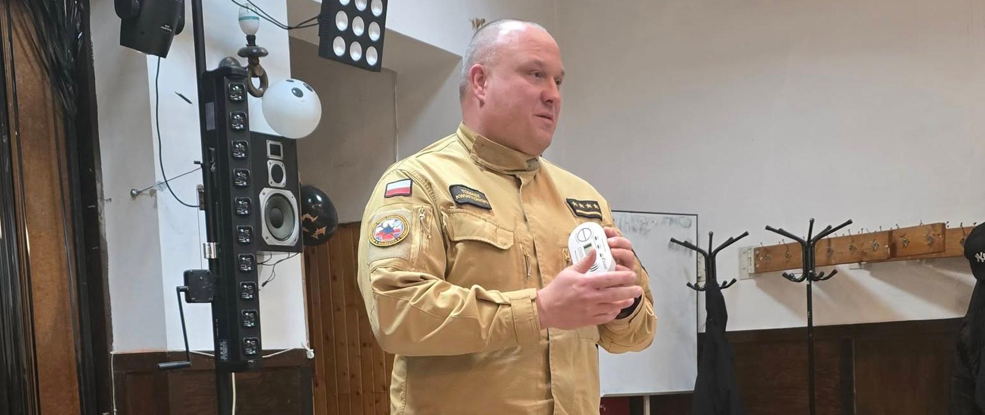 Strażak stoi w sali wyposażonej w profesjonalny sprzęt oświetleniowy i nagłośnieniowy. Trzyma w rękach czujkę z wyświetlaczem i przemawia, mając przed sobą mikrofon leżący na stole.