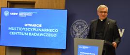 Mężczyzna w czarnym garniturze z koloratką stoi przy mównicy, za nim ekran z napisem Otwarcie Multidyscyplinarnego Centrum Badawczego.