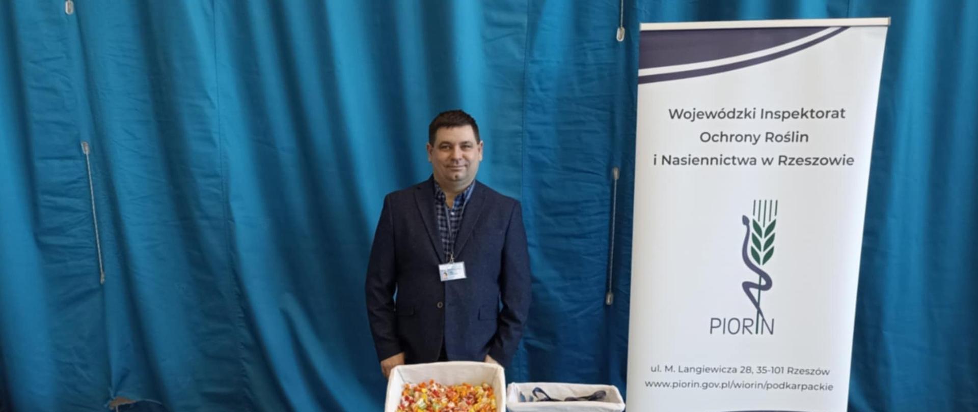 Mężczyzna ubrany w granatowy garnitur stoi przed stołem na którym leżą poukładane kolorowe ulotki, koszyk ze słodyczami oraz gadżety- smycze i długopisy. W tle za mężczyzna wisi duża niebieska kotara a po prawej stronie stoi biało granatowy baner z logiem instytucji. Podłoga w pomieszczeniu jest ciemna.