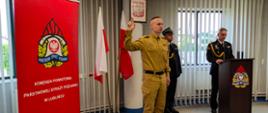 Zdjęcie zrobione w pomieszczeniu strażnicy na tle godła Rzeczpospolitej Polskiej, dwóch Flag Rzeczpospolitej Polskiej oraz po lewej stronie banneru Komendy Powiatowej PSP w Lublińcu. Na pierwszym planie nowoprzyjęty strażak stojący na baczność i ubrany w umundurowanie służbowe składa ślubowanie podnosząc prawą rękę, po prawej stronie Komendant w ubraniu galowym stojący za mównicą oraz dowódca uroczystości w umundurowaniu galowym z pasem i w czapce.