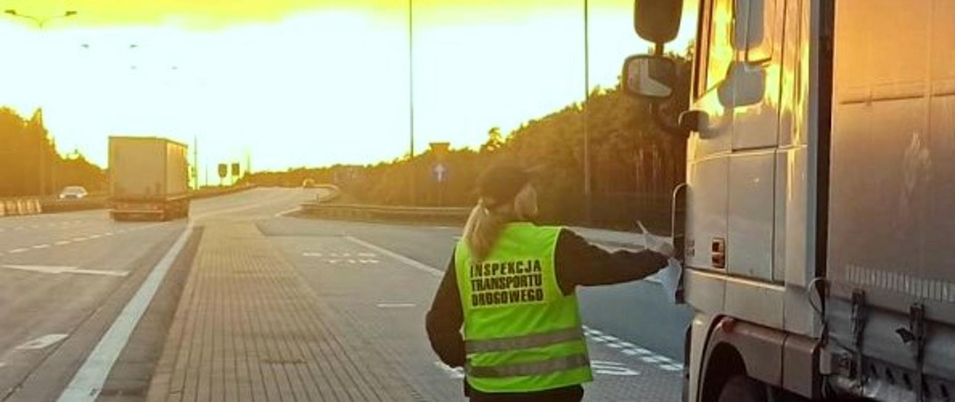 Inspektorka w kamizelce odblaskowej stoi obok białej kabiny ciągnika siodłowego. W tle wschód słońca.