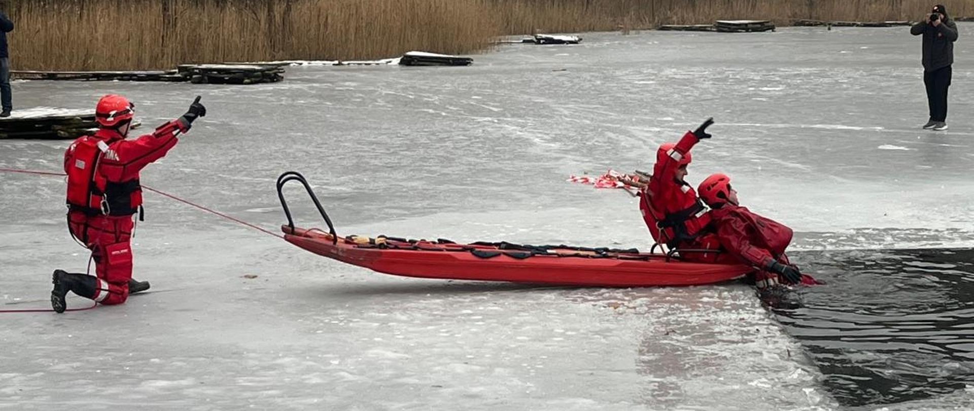 Strażacy na zamarzniętym jeziorze z wykorzystaniem sani lodowych prezentują jak wyciągają poszkodowanego z wody