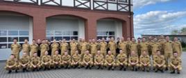 Grupowe zdjęcie kilkudziesięciu strażaków w mundurach służbowych w kolorze piaskowym. W tle budynek.