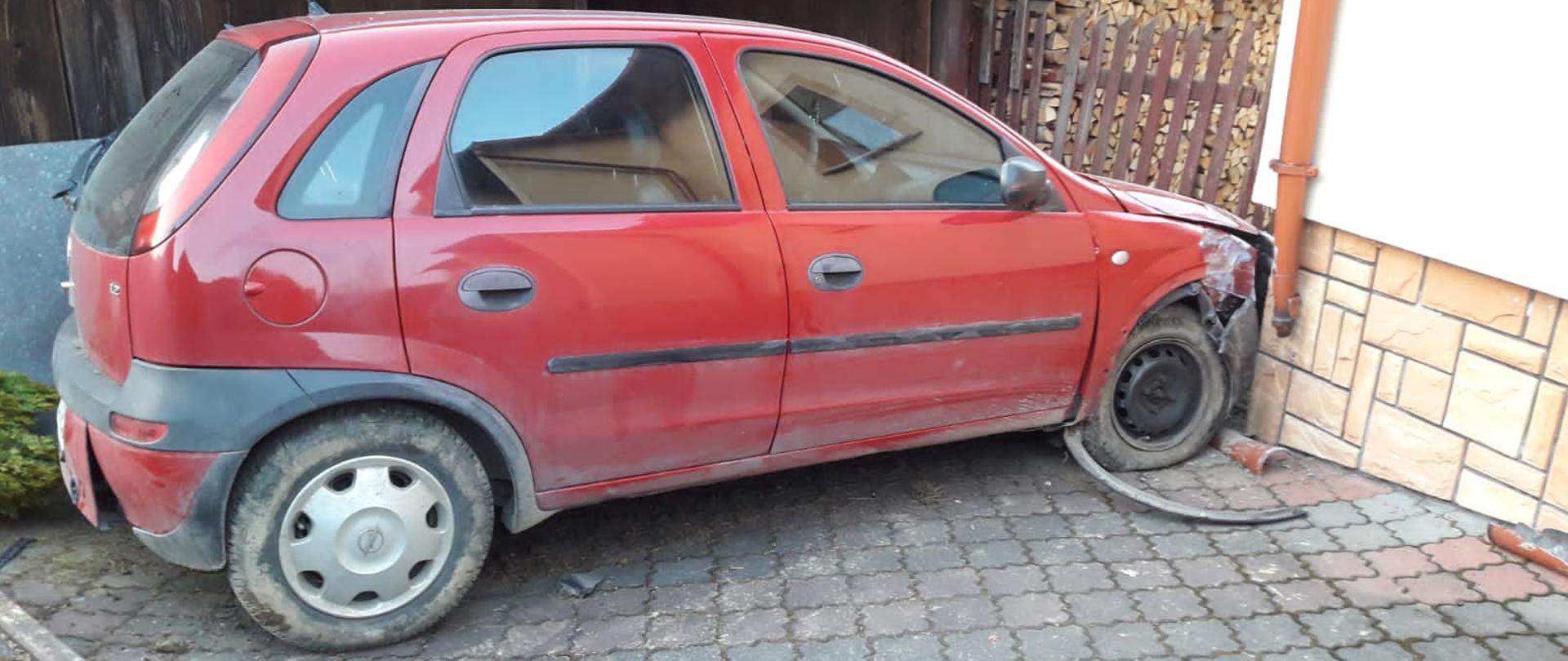 Samochód osobowy koloru czerwonego, przodem przy narożniku budynku. 