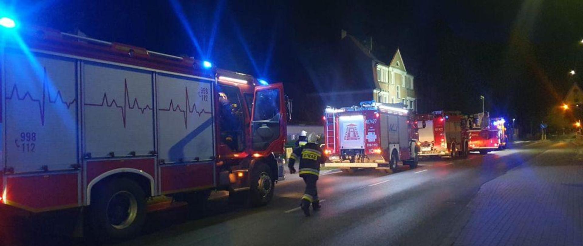 Zdjęcie wykonane w nocy. Całość oświetlona lampami błyskowymi wozów gaśniczych. Wzdłuż drogi stoją pojazdy gaśnicze. W tle budynek objęty działaniami.