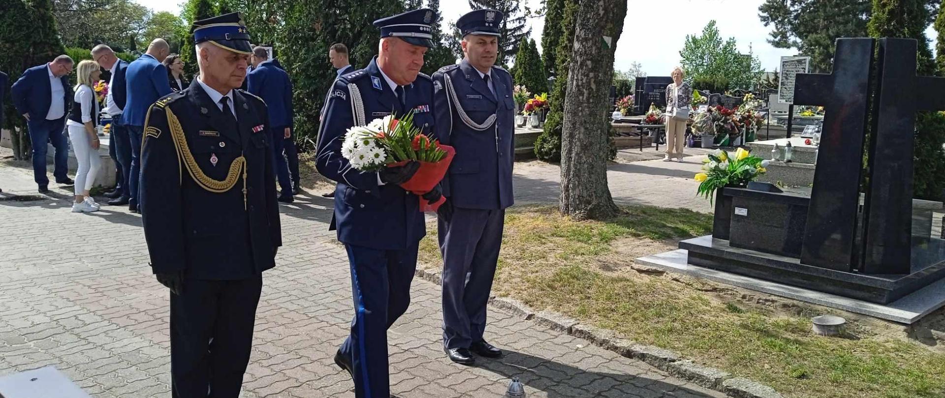 Rawicki cmentarz parafialny. Delegacja służb mundurowych: PSP, Policji i Służby Więziennej maszeruje przed krzyż, aby złożyć wiązankę kwiatów. Kwaity trzyma w rękach policjant. Wszyscy funkcjonariusze występują w mundurach wyjściowych ze sznurem. W tle pozostali uczestnicy uroczystości, a także nagrobki i drzewa.