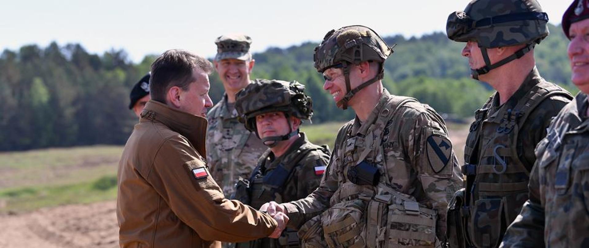The head of the Ministry of National Defence greets US and Polish soldiers at the Drawsko training ground. In the background forest and sand. Soldiers in a row standing in front of the Minister of Defence.
