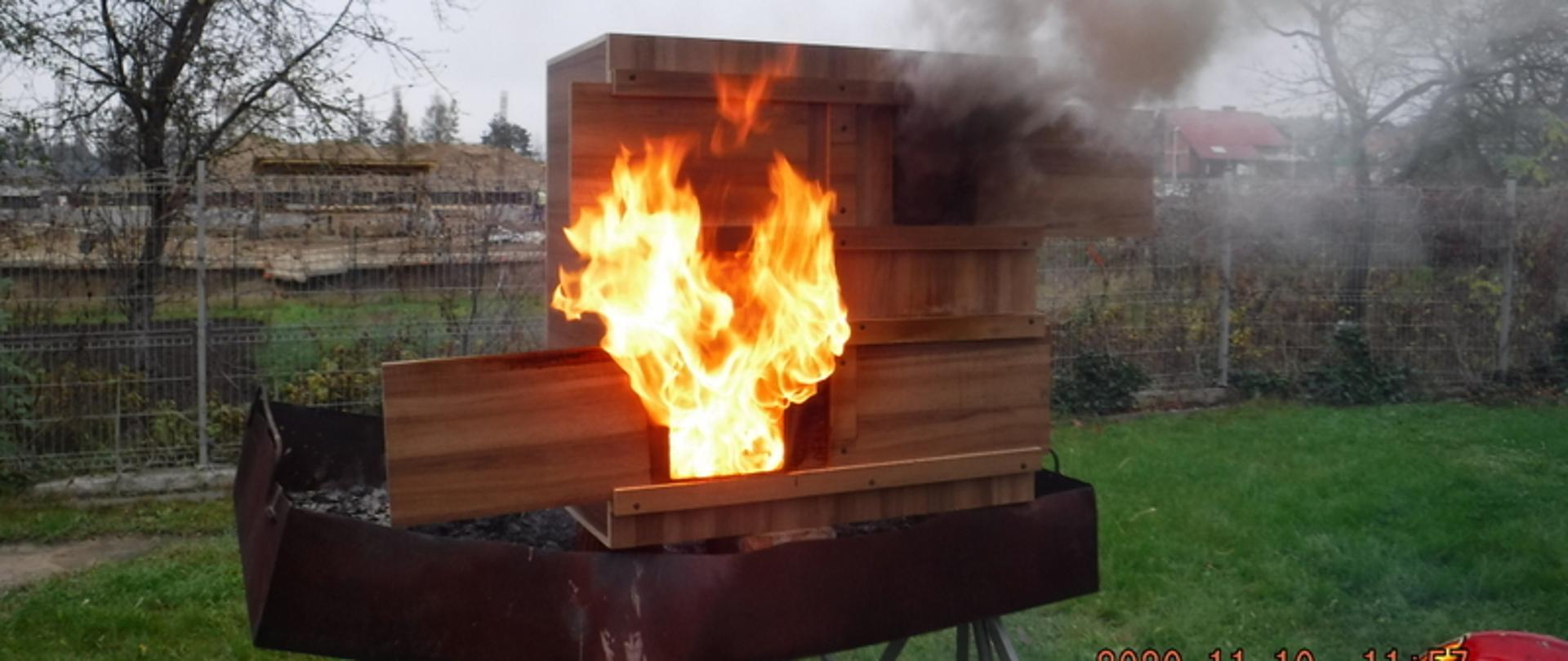 Zdjęcie główne przedstawia płonącą makietę budynku. Z okna na parterze buchają natomiast z okna na piętrze wydobywa się szary dym