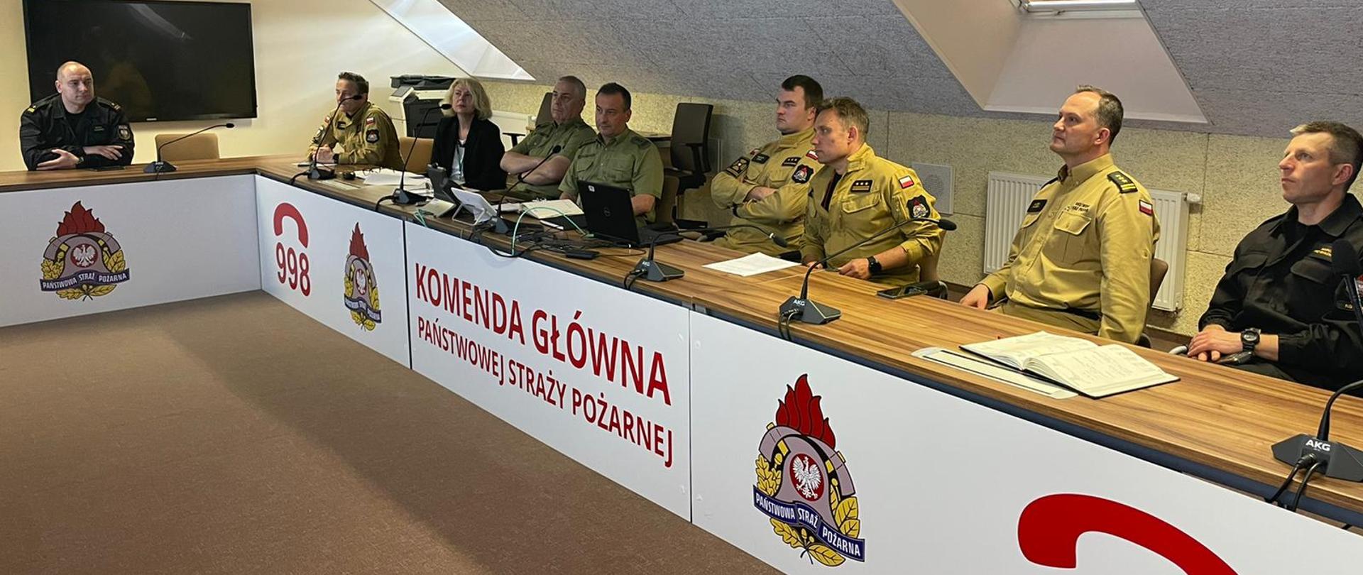 Spotkanie robocze. Kobieta i ośmiu mężczyzn siedzący przy stole. Patrzą przed siebie. 