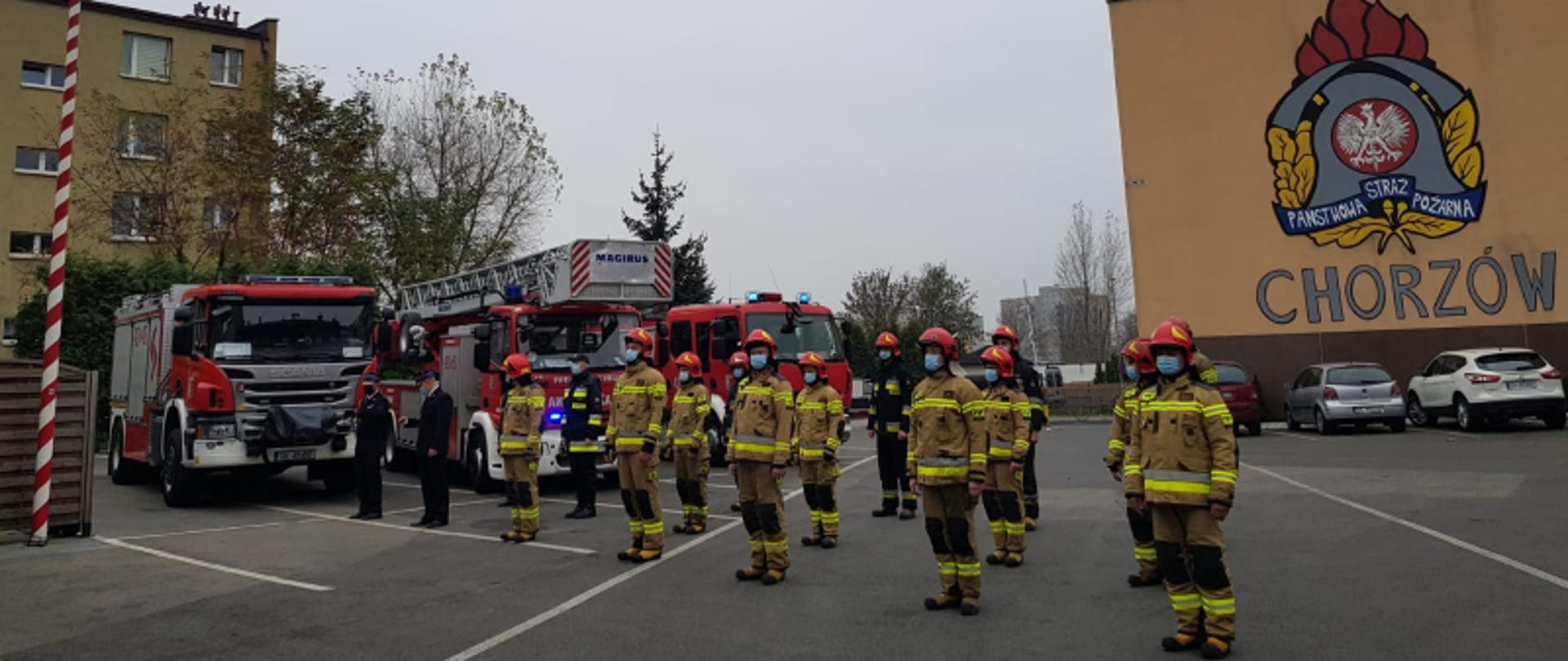 Na fotografii widać strażaków ubranych w ubrania bojowe oraz Komendanta Miejskiego stojących w szyku na placu wewnętrznym Komendy Miejskiej Państwowej Straży Pożarnej w Chorzowie. Strażacy śpiewają hymn państwowy przy podniesionej fladze biało - czerwonej. Obok szyku strażaków stoją dwa samochody gaśnicze oraz drabina mechaniczna. W tle na murze budynku widnieje mural - logotyp Państwowej Straży Pożarnej.