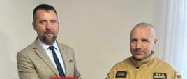 Po lewej stronie stoi mężczyzna w garniturze, z teczką w ręku z herbem strażackim oraz logo ZOSP RP (Związek Ochotniczych Straży Pożarnych RP). Po prawej stronie znajduje się strażak w piaskowym mundurze z emblematami i stopniami służbowymi. Na jego mundurze widoczna jest naszywka z nazwiskiem oraz polska flaga. On również trzyma identyczną teczkę. Tło stanowi biuro z zegarem na ścianie, roślinami w doniczkach i firanką w oknie. Zdjęcie dokumentuje ważny moment – prawdopodobnie podpisanie umowy, porozumienia lub wręczenie nominacji związanej z działalnością strażacką. Atmosfera jest oficjalna, ale przyjazna.