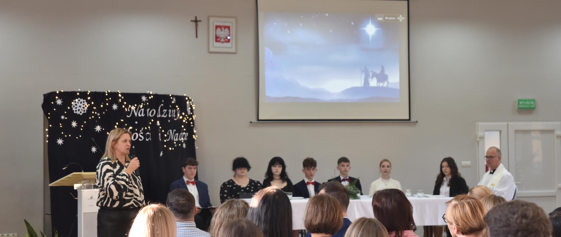 Na zdjęciu widoczna scena, na niej grupa młodzież i ksiądz siedzący przy wigilijnym stole, w tle ekran z widocznymi postaciami Józefa i Maryi na osiołku, po lewej stronie sali pani dyrektor przemawiająca do społeczności szkolnej.