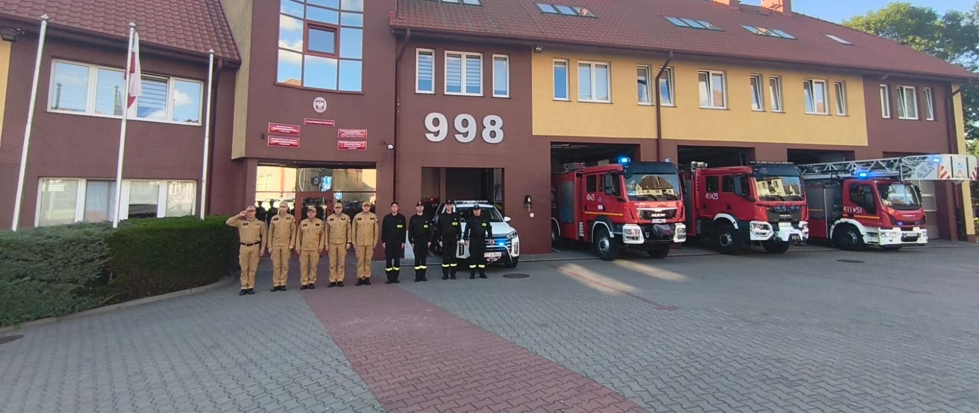 Szeroki kadr na budynek Komendy Powiatowej PSP z widocznymi garażami i zaparkowanymi samochodami ratowniczo-gaśniczymi. Strażacy ustawieni w szeregu przed jednostką.