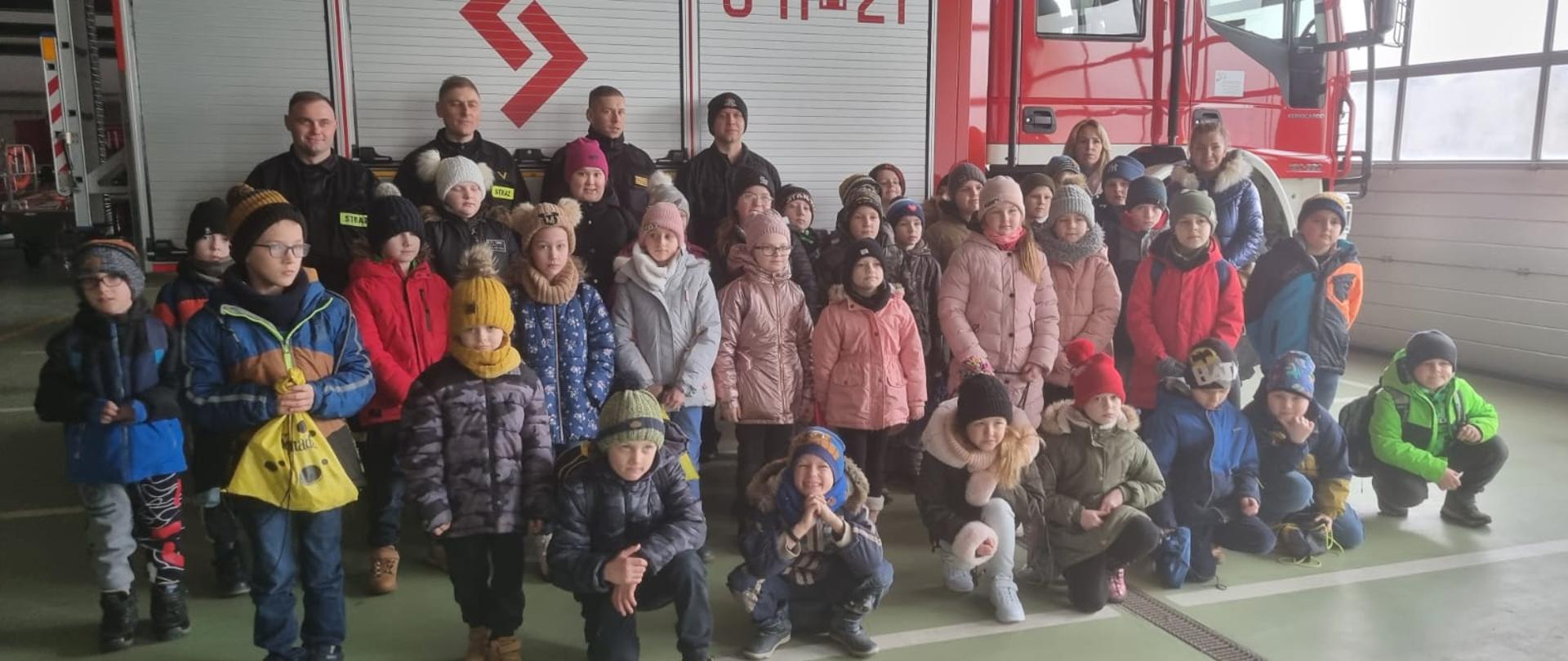 Wizyta w ramach akcji „Odwiedź strażnicę i salę edukacyjną typu Ognik” w KP PSP w Sokołowie Podlaskim + na zdjęciu dzieci i strażacy stoją w garażu JRG.
