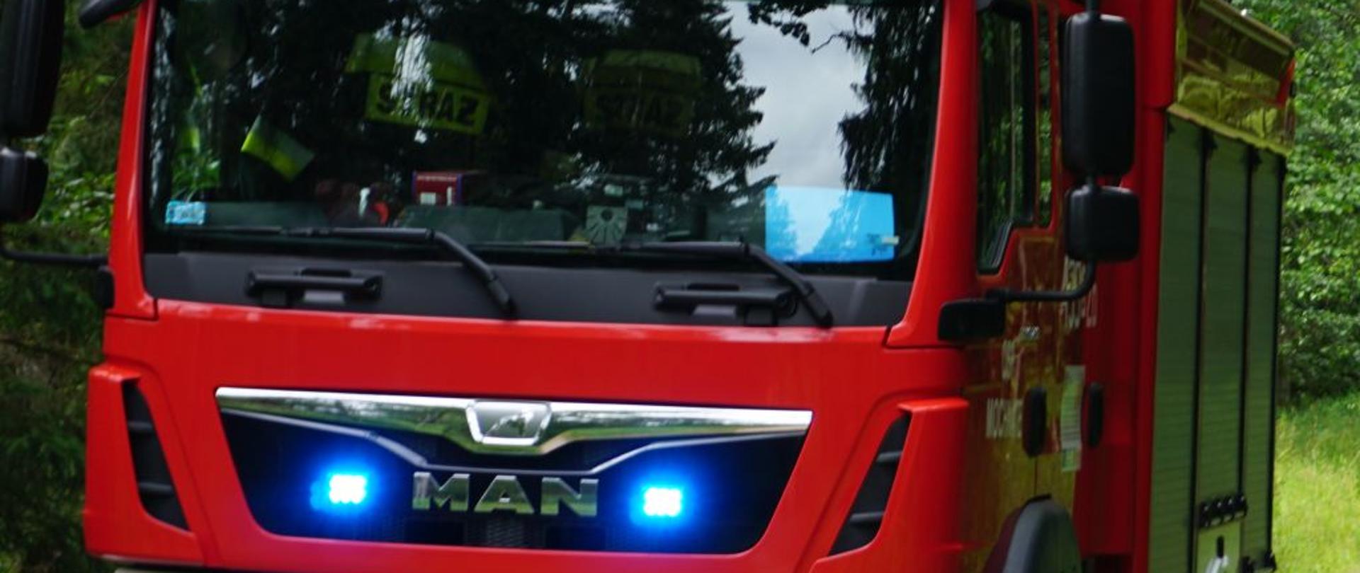 Na tle zielonych drzew na sygnałach jedzie czerwony strażacki wóz bojowy.