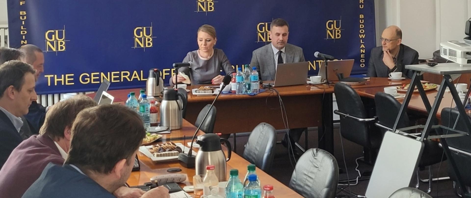 Uczestnicy konferencji siedzący przy stole z laptopami i napojami; w tle baner „GUNB”, na stole projektor, kawa, woda i przekąski.