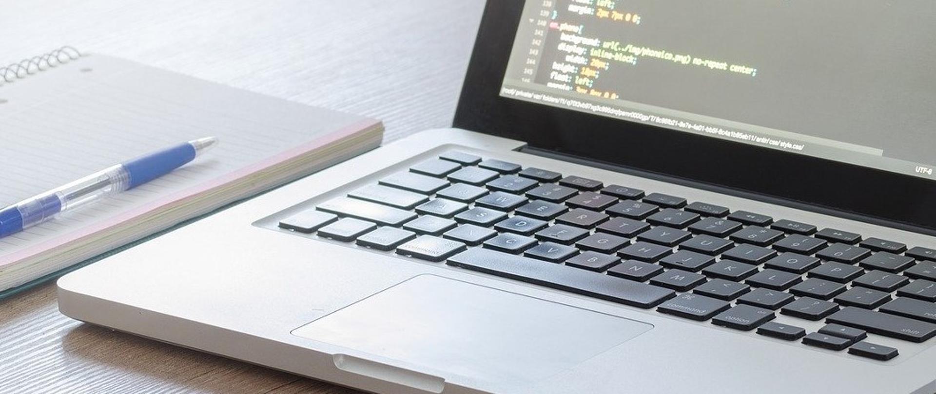 Na biurku otwarty laptop, obok notes i długopis