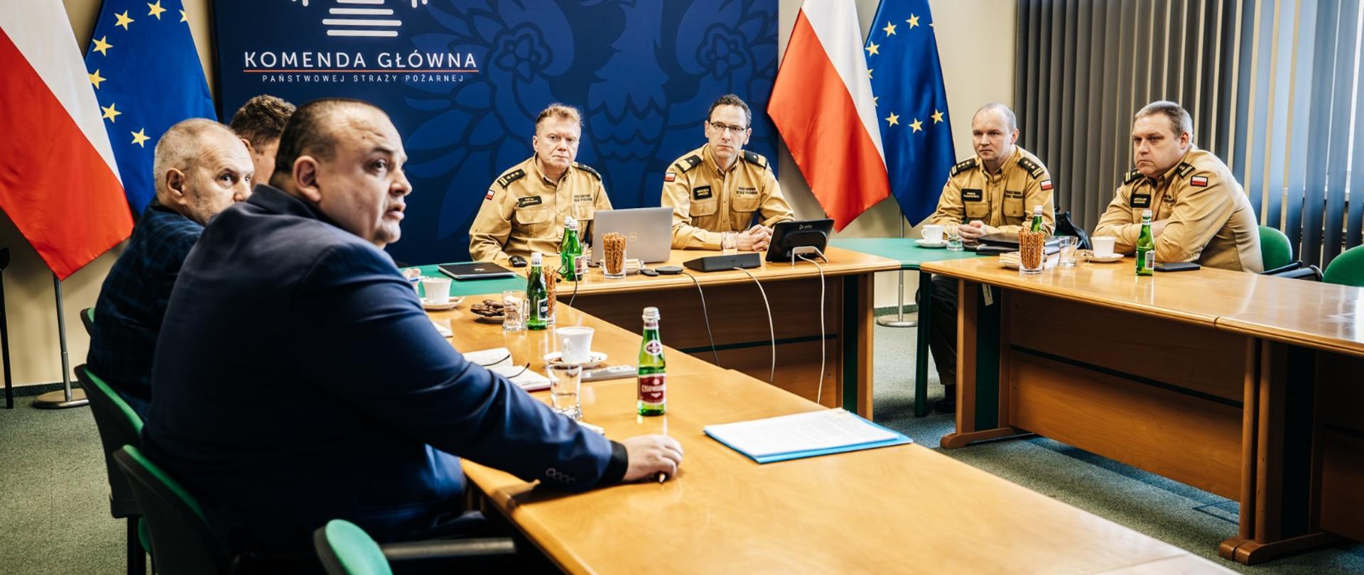 Sala konferencyjna Komendy Głównej Państwowej Straży Pożarnej. Przy stołach ustawionych w podkowę siedzą przedstawiciele kierownictwa PSP w umundurowaniu służbowym oraz reprezentanci strony związkowej w garniturach. W tle widoczne są flagi Polski i Unii Europejskiej oraz tablica z godłem i nazwą Komendy Głównej PSP. Na stołach znajdują się dokumenty, laptopy, mikrofony oraz napoje.
