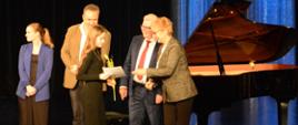na zdjęciu w sali widowiskowej JDK, podczas koncertu laureatów widnieje uczennica PSM I stopnia w Jaśle, która obiera dyplom od jury. Kolorystyka zdjęcia jest niebiesko-biało-brązowo-czarna.