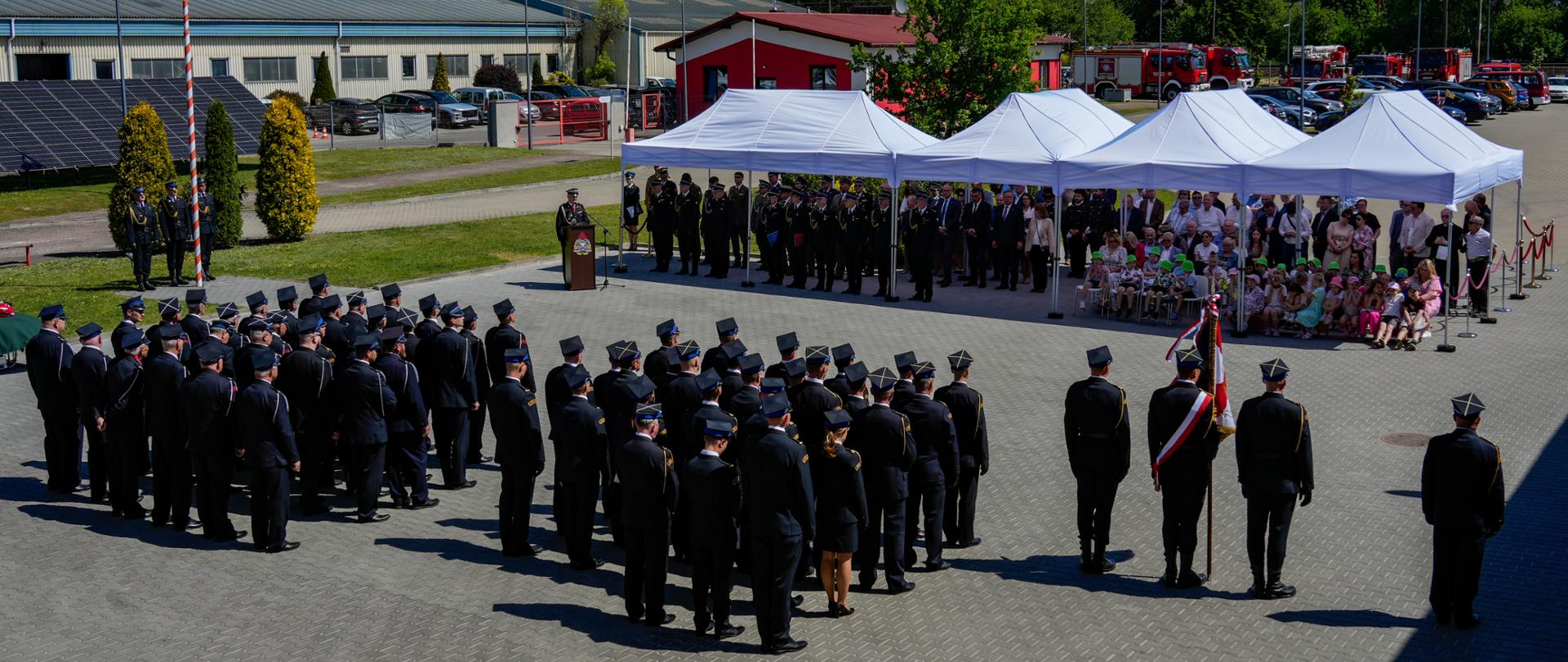 Na zdjęciu widać uroczystą zbiórkę strażacką odbywającą się na placu przed remizą. Kilkudziesięciu strażaków w galowych mundurach stoi w zwartym szyku obok masztu z biało-czerwoną flagą. Naprzeciw nich, pod białymi namiotami, siedzą i stoją zaproszeni goście oraz rodziny z dziećmi. W tle widoczne są wozy strażackie, budynki straży pożarnej oraz las. Zdjęcie wykonano w słoneczny dzień z podwyższonej perspektywy.