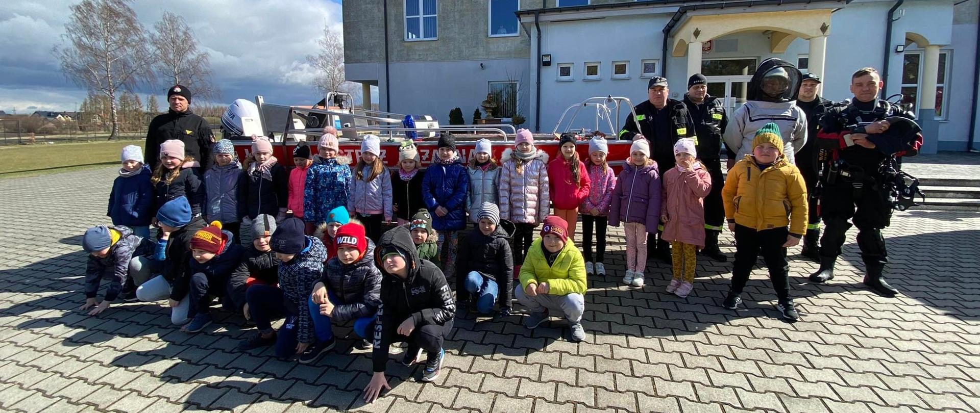 Dzieci oraz Strażacy stoją przed budynkiem szkoły przy strażackiej łódce.