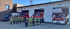 Fotografia przedstawia strażaków w ubraniach bojowych z hełmami na głowach stojących w szeregu w trakcie uroczystego uczczenia minutą ciszy strażaków poległych na Ukrainie na dalszym planie widać pojazdy pożarnicze oraz budynek jednostki ratowniczo gaśniczej