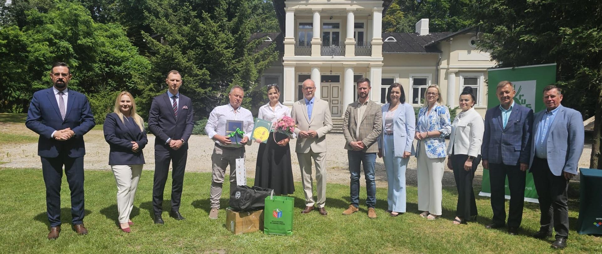 Na trawniku przed pałacem w Borowicach stoi grupa uczestników i organizatorów wydarzenia. W centrum znajdują się laureaci z dyplomem, nagrodami i kwiatami, otoczeni przez przedstawicieli KRUS oraz zaproszonych gości. W tle widoczny jest zabytkowy pałac i zieleń parkowa.