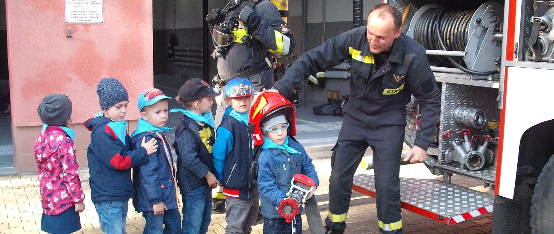 Chłopiec w hełmie strażackim trzyma w rękach prądownicę z podpiętym wężem. Obok stoi strażak i podtrzymuje hełm na głowie chłopca. Za chłopcem kolejka dzieci.